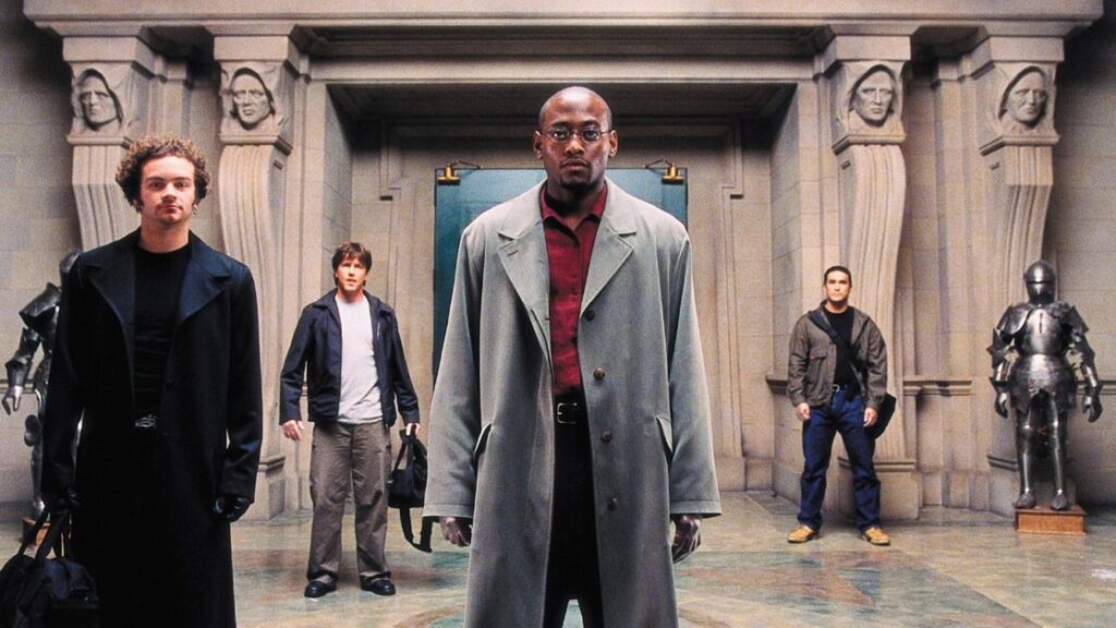 Four men stand in an ornate hallway with statues and armored suits in the background, facing the camera. The central figure is wearing a long coat and glasses.