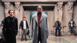 Four men stand in an ornate hallway with statues and armored suits in the background, facing the camera. The central figure is wearing a long coat and glasses.