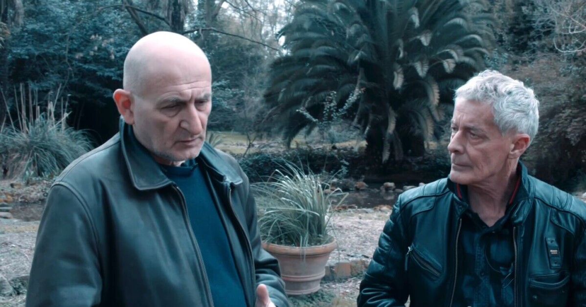 Two men stand outdoors in a garden-like setting with greenery in the background. Both are wearing dark jackets and are engaged in a conversation.