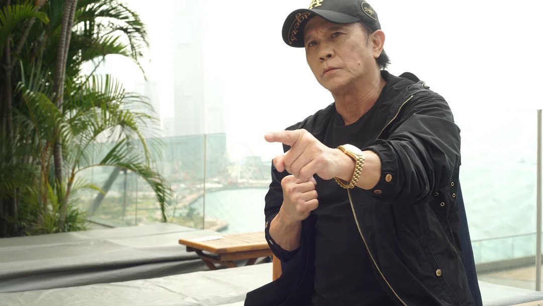 A man in a black jacket and cap makes a pointing gesture while sitting outdoors near a glass barrier with greenery and a body of water in the background.