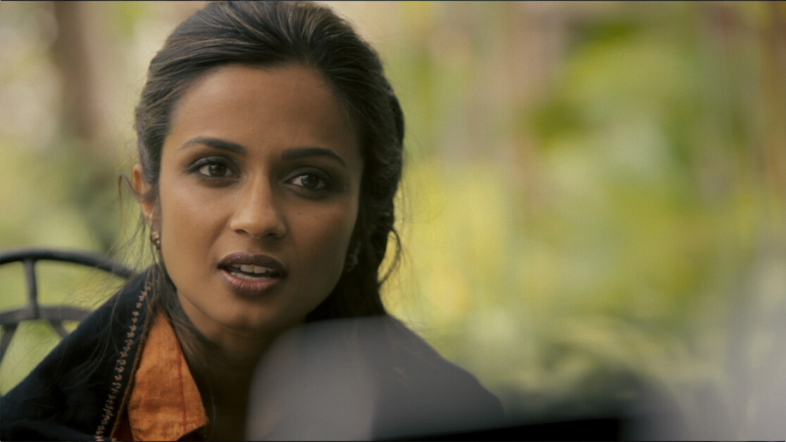 A woman with dark hair and a serious expression is seen in a close-up shot, wearing a dark jacket and an orange collar shirt. The background is blurred with greenery.