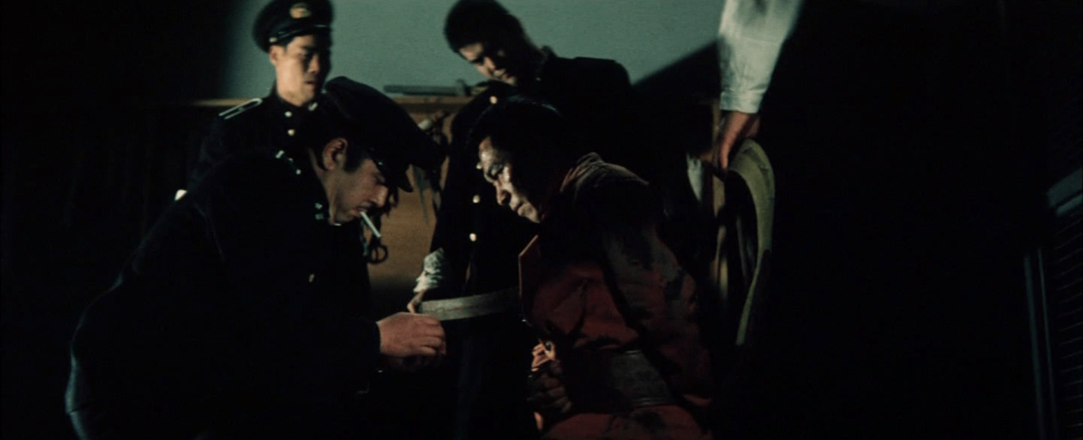 Group of men, some in uniforms, surrounding and interacting with a person sitting in a chair in a dimly lit room.
