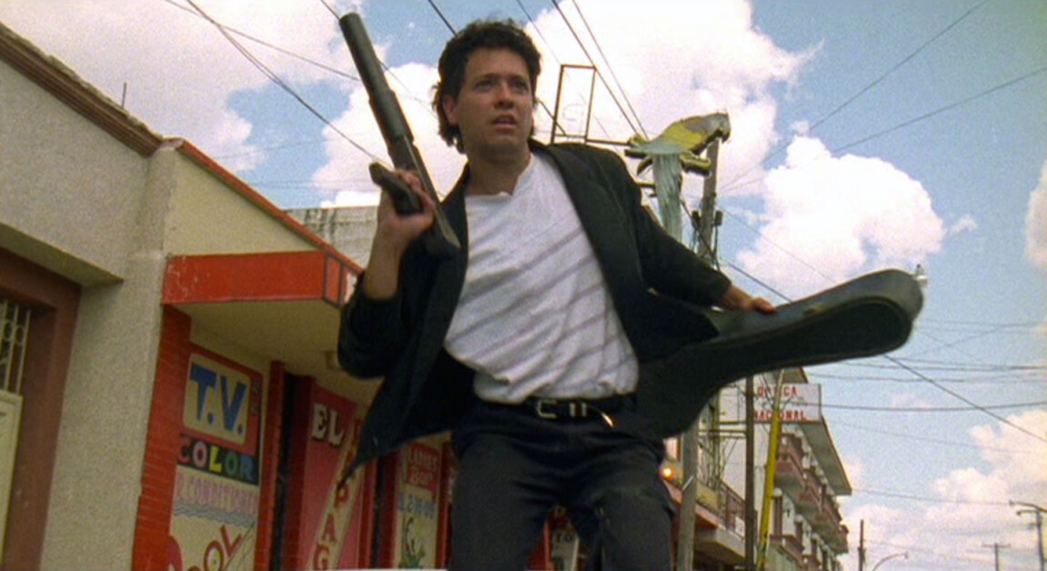 A man holding a shotgun and guitar case stands on top of a moving vehicle in an urban area with buildings and wires visible in the background.