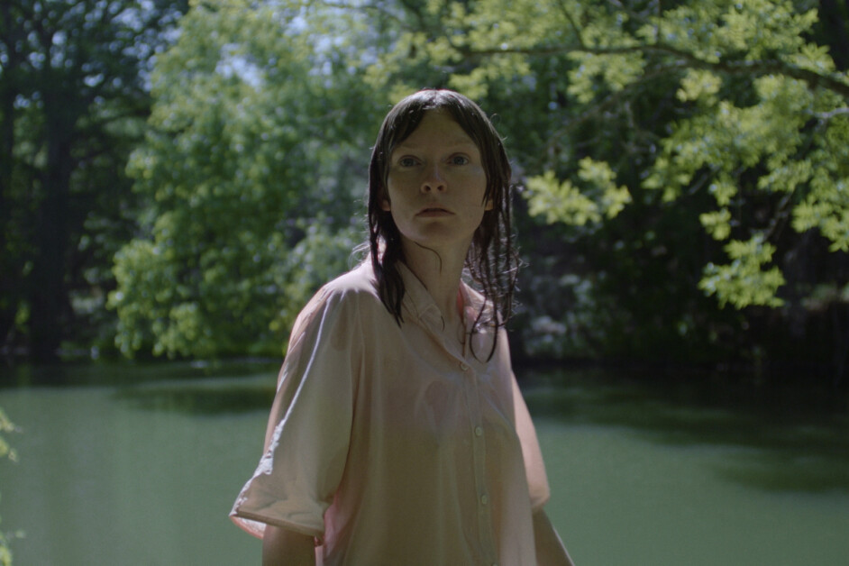 A person with wet hair stands outdoors by a body of water, wearing a light-colored shirt, and surrounded by green trees in the background.