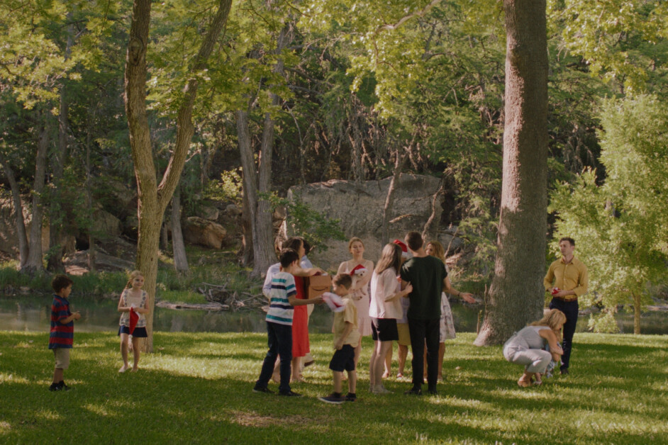 A group of children and adults are gathered in a grassy park area with trees and a river in the background. Some are interacting and playing while others stand or sit on the grass.