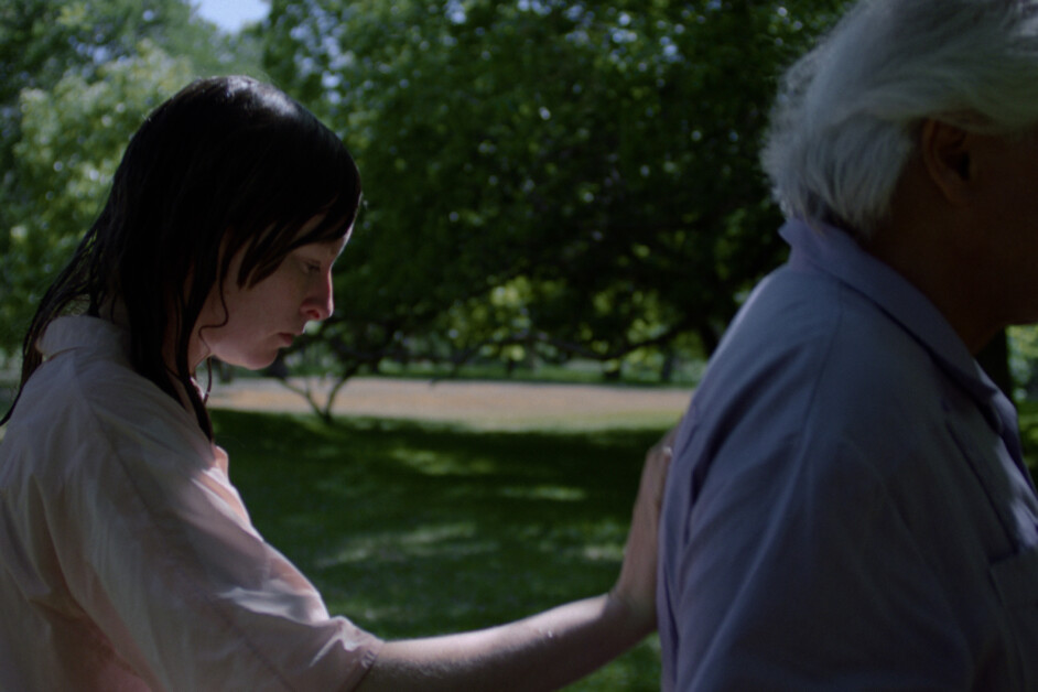 A person with wet hair touches the back of an older person with gray hair in a park setting.