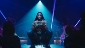 A performer dressed in a sparkling outfit and thigh-high boots poses dramatically on a dimly lit stage with blue lighting, surrounded by an audience.