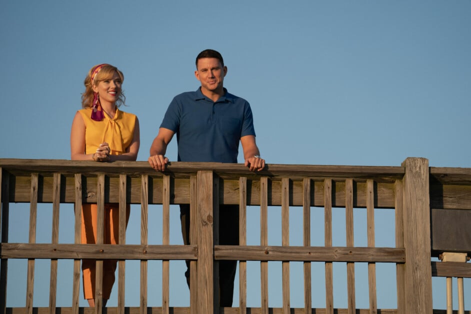 A woman and a man stand on a wooden platform, leaning on the railing. The sky is clear and blue.
