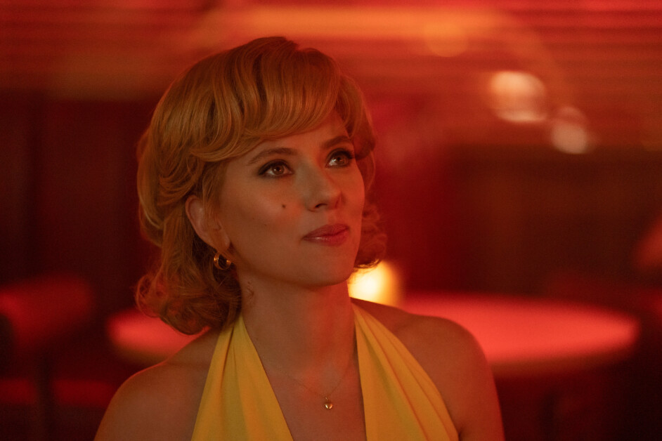 A woman with short, wavy hair glances upward in a dimly lit room. She wears a yellow halter top and a necklace. The background is shaded in red tones.