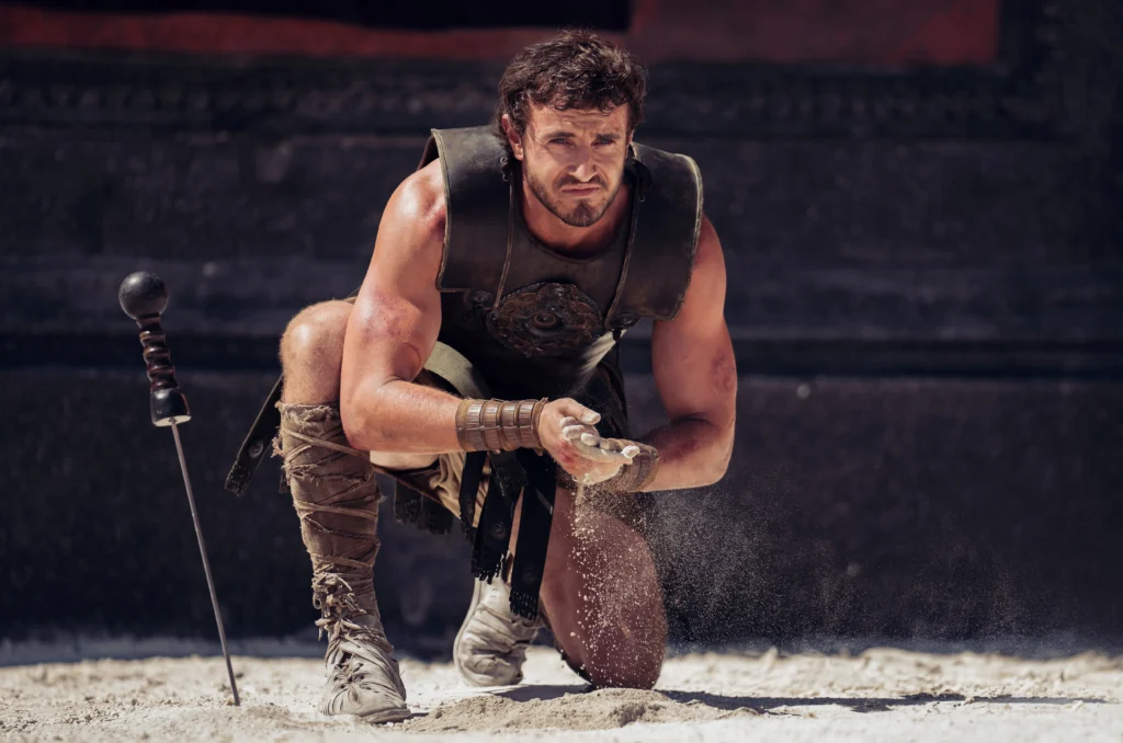 A person in Roman soldier attire kneels on the ground, holding sand in one hand while the other hand rests on the ground. A sword is stuck in the sand nearby.