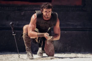 A person in Roman soldier attire kneels on the ground, holding sand in one hand while the other hand rests on the ground. A sword is stuck in the sand nearby.