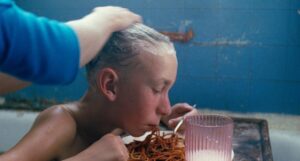 A person with whitened hair sits in a bathtub, bending over a plate of spaghetti with a hand resting on their head. A pink tumbler is next to the plate.
