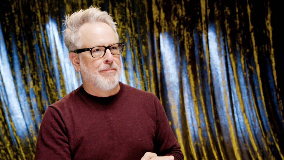 A man with gray hair, glasses, and a beard, wearing a maroon sweater, stands against a background with vertical blue and yellow streaks.
