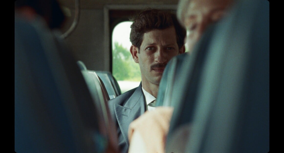 A man with a mustache, wearing a suit and tie, sits on a bus looking forward. The seatbacks and partial view of another passenger are seen in the foreground.