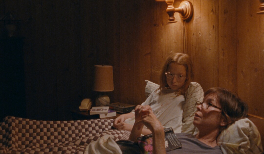 Two women are lying in bed under a quilted blanket with wooden wall paneling and reading lamps behind them; one is reading a book while the other is looking intently at her.