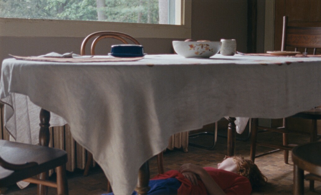A person lying on the floor under a table with a white tablecloth, various dishes on the table, and a window in the background.