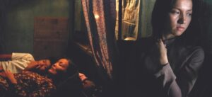 A woman stands pensively in the foreground while two people lie down together in a dimly lit room in the background.