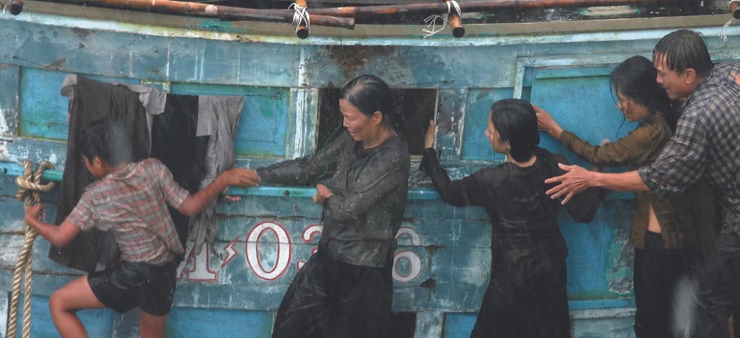 A group of people are on an old, blue wooden boat in the rain. Some are holding onto the boat while others appear to be looking or reaching out. Clothing is hung to dry on the side of the boat.