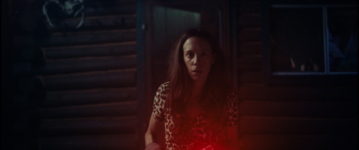 A woman wearing a leopard print shirt looks startled while standing in front of a log cabin at night. A red light is illuminating her face.