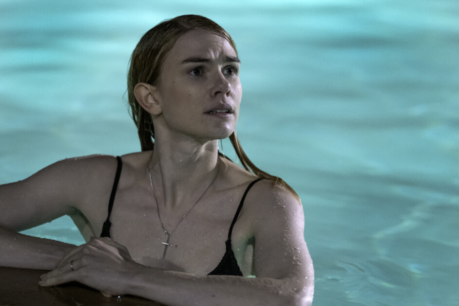 A person with wet hair and a serious expression stands in a swimming pool, holding onto the pool edge while wearing a necklace with a cross pendant.