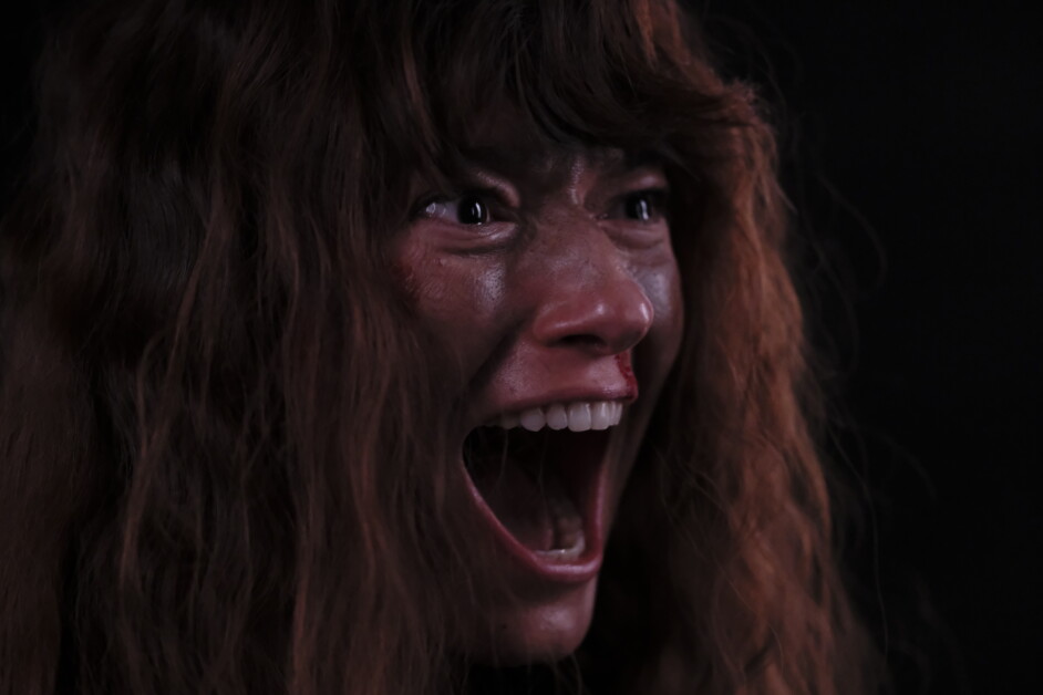 A person with long hair is shown in close-up, screaming with a wide open mouth and visible tension on their face, against a dark background.