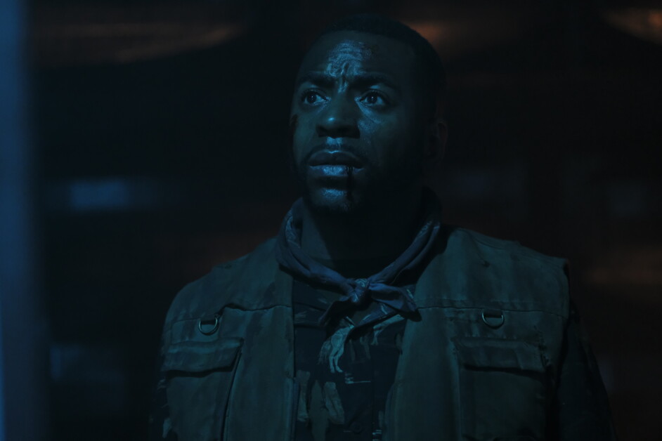 A man in dark clothing with a serious expression looks upward. The lighting is dim and blue-toned, creating a dramatic atmosphere.