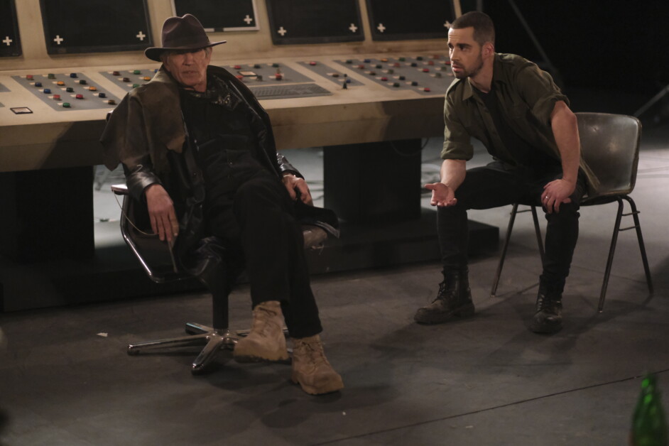 Two men are sitting in front of a large console with various buttons and meters. One man is wearing a hat and coat, sitting in a relaxed manner, while the other man, in casual attire, appears engaged.