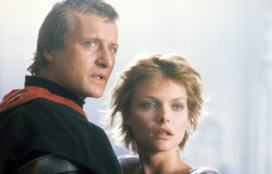 ‘Ladyhawke’ Blu-Ray Review – Cursed Lovers Fight For One Another In Epic Fantasy Adventure Two individuals with serious expressions. The person on the left wears a black outfit with a red detail, while the person on the right has short blonde hair and a white top. Misty background.