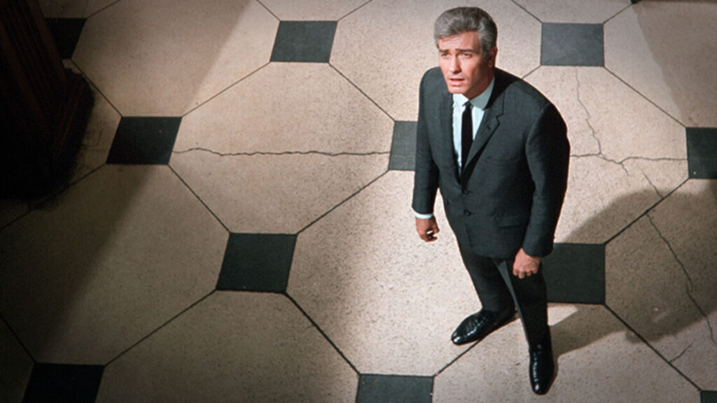 A man in a suit stands on a checkered floor, looking upwards.