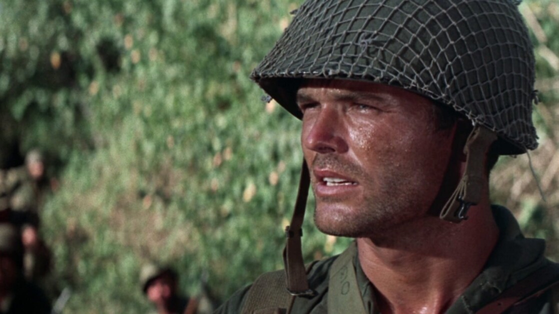 A soldier with a weary expression stands outdoors wearing a helmet and military uniform, with foliage visible in the background.