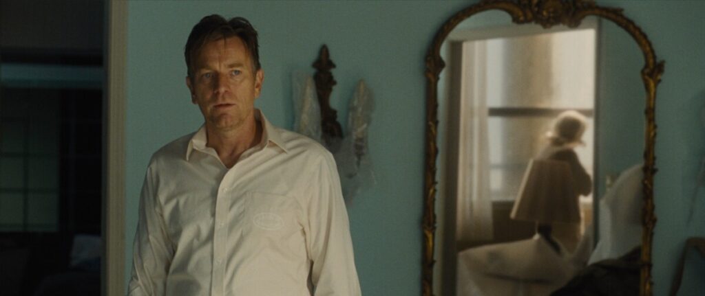 A man in a white shirt stands near a wall-mounted mirror reflecting a woman dressed in white, looking out of a window in a dimly lit room.