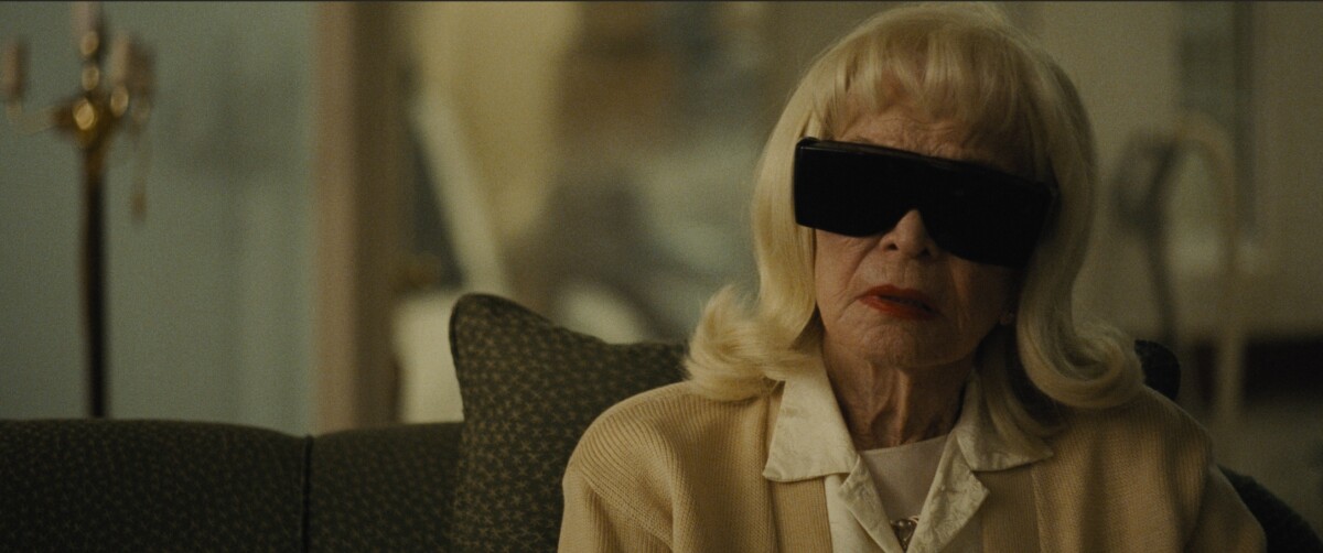 An elderly woman with long blonde hair sits on a couch wearing large black sunglasses and a white outfit.