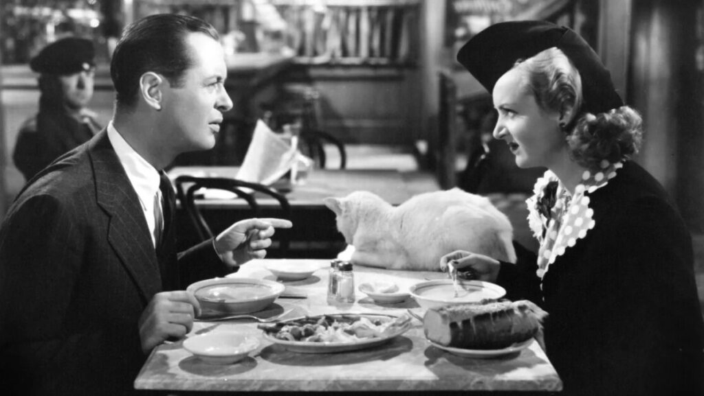 A man and a woman are seated at a table in a restaurant, eating a meal with a loaf of bread and a white cat lying on the table between them.