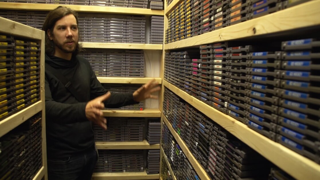 A person stands in a narrow aisle surrounded by shelves filled with numerous video game cartridges. They are gesturing with their hands while appearing to explain something.