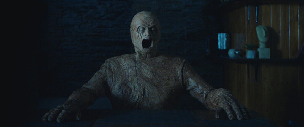 A humanoid figure with a textured, tree-like appearance sits at a table, mouth open, in a dimly lit room with wooden shelves and a stone wall background.