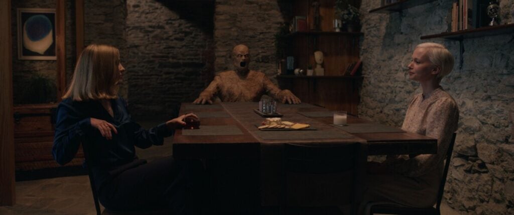 Two women sit opposite each other at a dining table with snacks. A mannequin is seated at the far end of the table. The room has stone walls, bookshelves, and framed artwork.