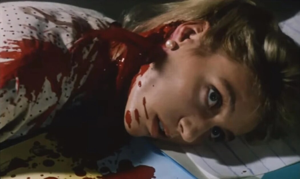 Person with long hair and earrings lying down, covered in blood, with eyes open in a scene from a horror movie.