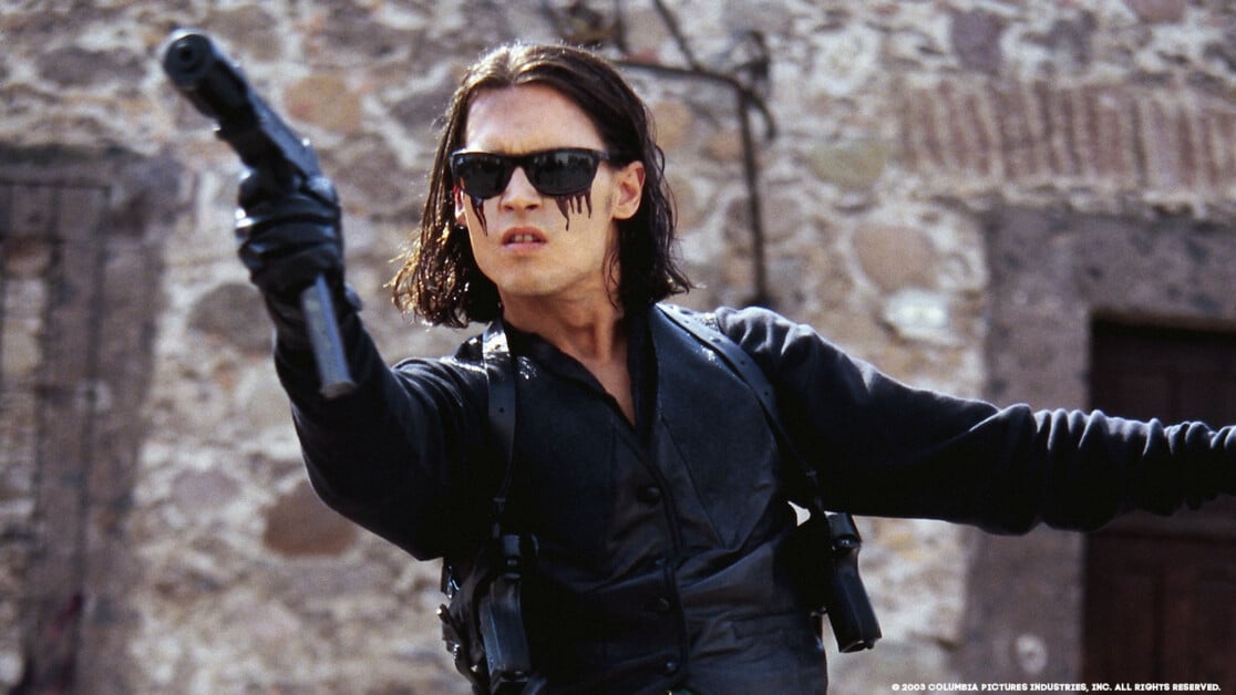 Person wearing sunglasses and dark clothing aims a gun with an intense expression. Stone wall in the background.