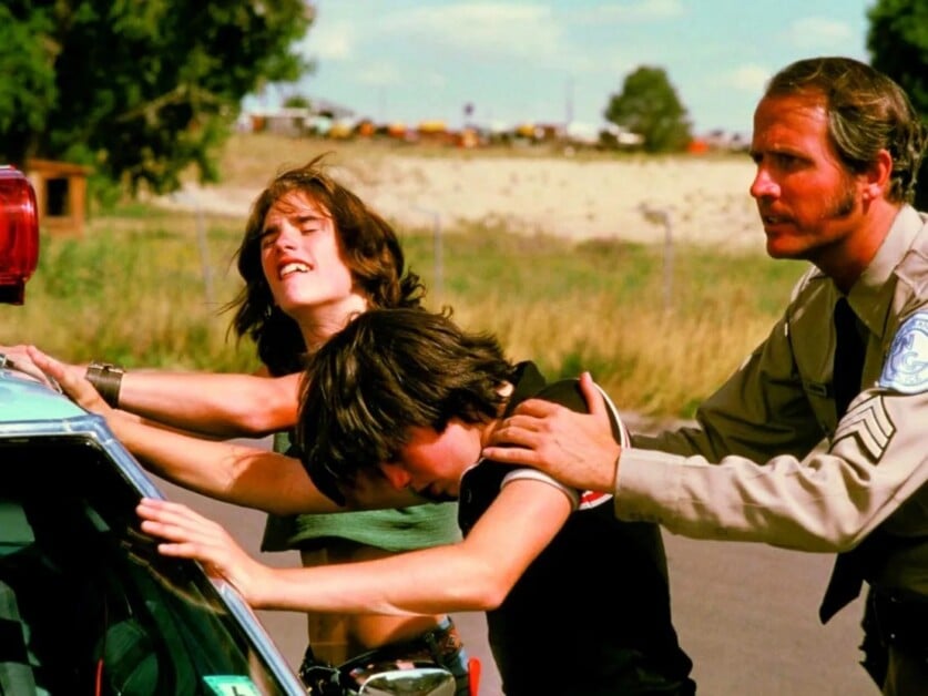 Two young individuals are being searched by a uniformed officer while leaning against a police car, with a rural landscape in the background.
