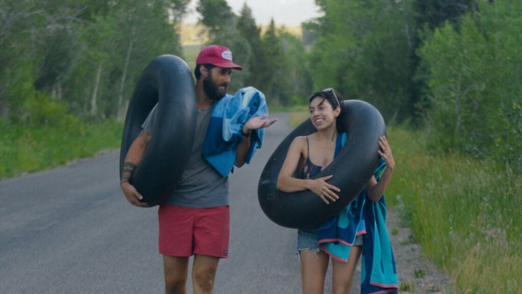 Two people are walking on a road surrounded by greenery, each carrying an inflatable tube and wearing casual summer clothes.