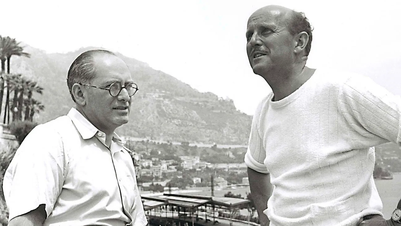 Two men are in conversation outdoors with a mountainous landscape in the background. One man is wearing a short-sleeve, buttoned shirt and glasses, while the other is wearing a light-colored sweater.