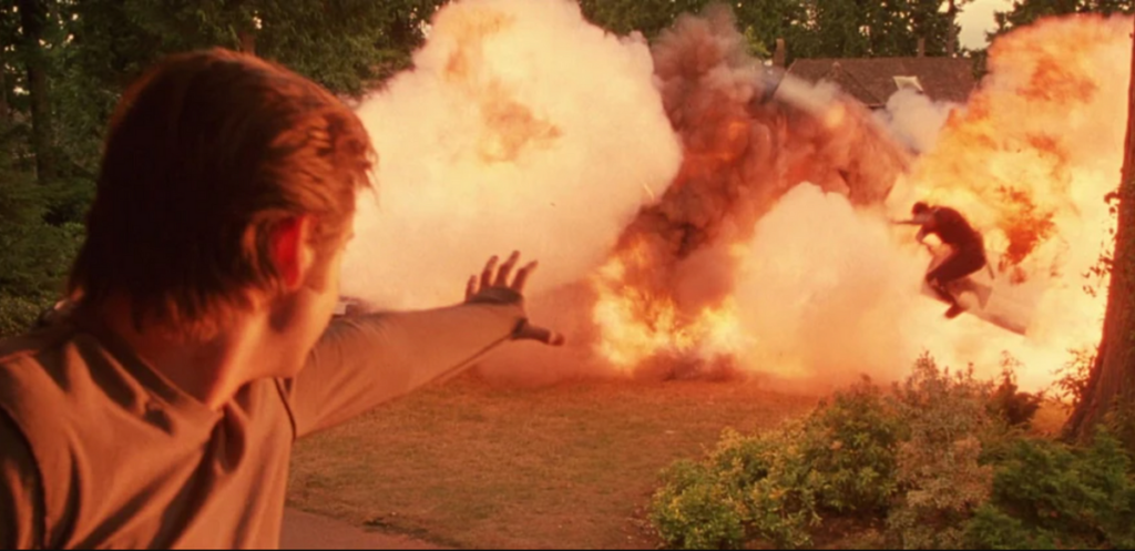 A person extends a hand towards a large explosion in an outdoor setting, while another person is being thrown by the blast.