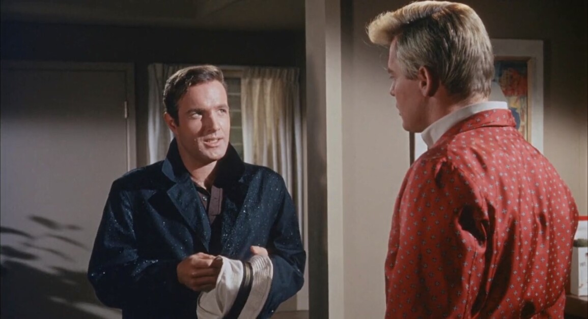 Two men standing indoors, facing each other. The man on the left holds a hat, while the man on the right wears a red robe with a pattern. They appear to be in conversation.