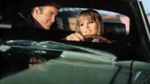 A man and a woman sit in a car. The woman is holding the steering wheel and appears to be talking, while the man looks at her with a slight smile.