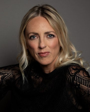A woman with long blonde hair is wearing a black lace top and earrings, looking directly at the camera with a neutral expression against a plain background.