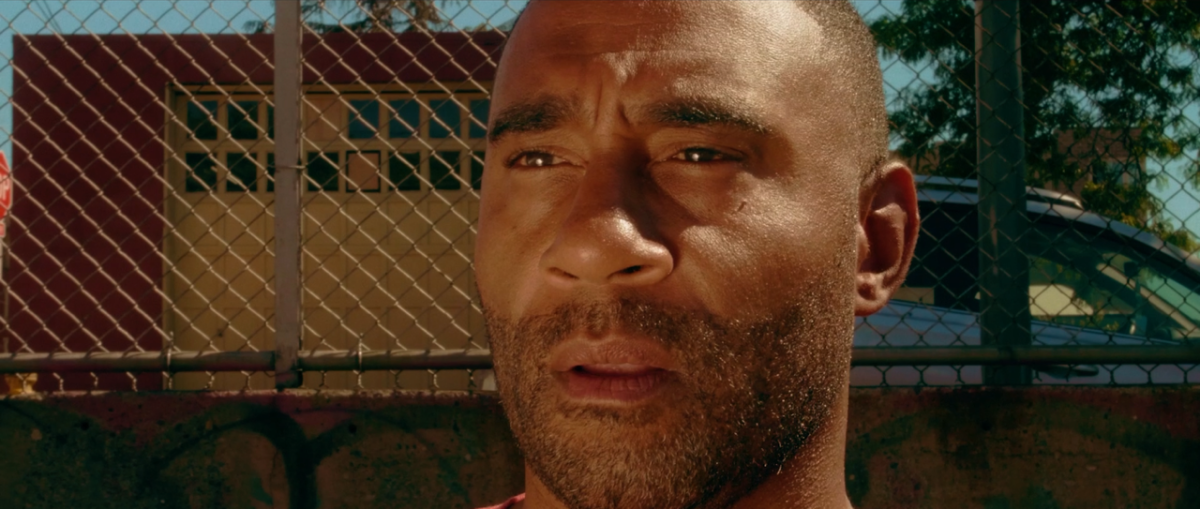 A close-up of a man's face with a concerned expression. He is standing outdoors in front of a chain-link fence and a building.