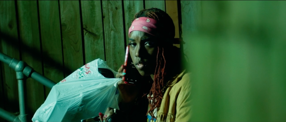 Person with a pink bandana, holding a phone to their ear, sitting against a wooden wall and holding a plastic bag. The lighting is dim and greenish.