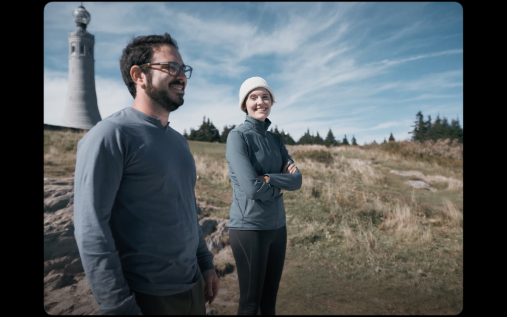 Two people wearing outdoor clothing stand on a grassy area near a lighthouse. One person is smiling with arms crossed, and the other is smiling with hands by their sides. The sky is partly cloudy.