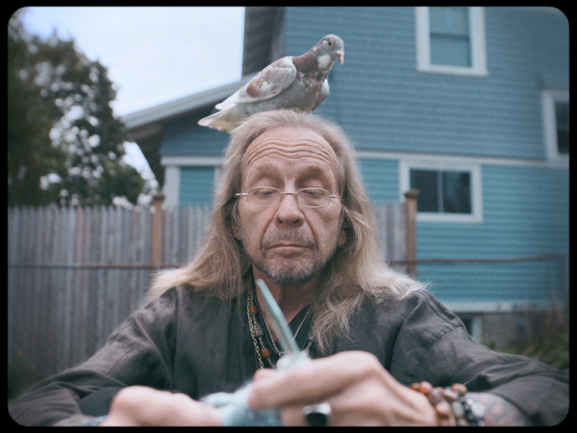 A man with long hair and glasses is sitting outdoors knitting, with a pigeon perched on his head. There is a blue house and wooden fence in the background.