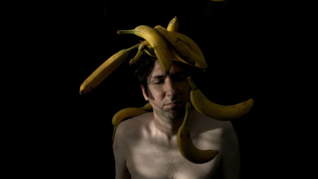 A shirtless person stands with eyes closed while multiple bananas are balanced on their head against a dark background.